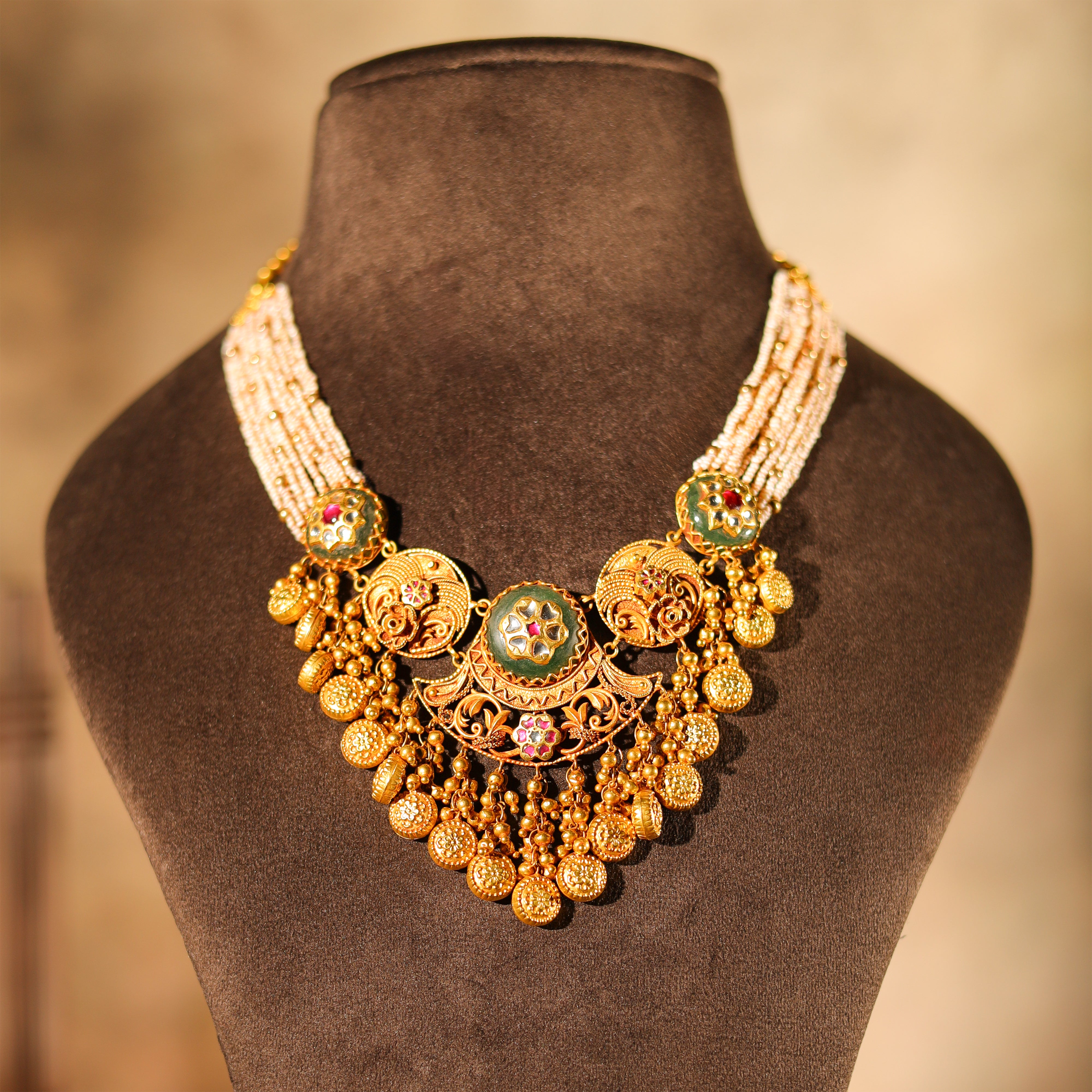 Ornate gold necklace with green and red stones on a brown mannequin bust.